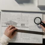 Hands holding a ruler and magnifying glass over architectural floor plans on a white table.